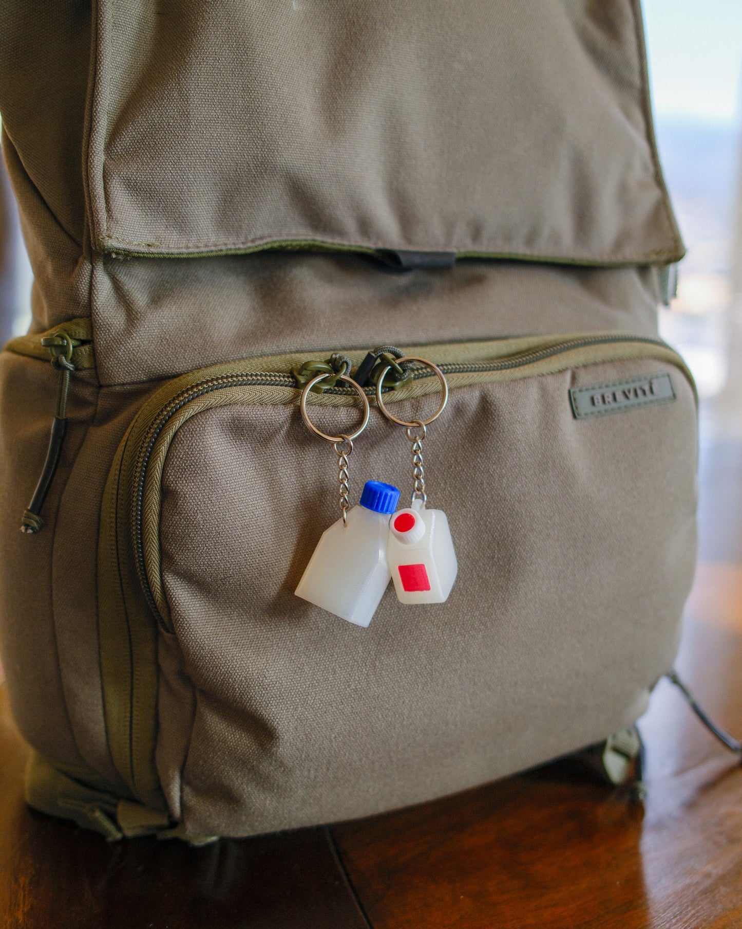 Clear cell culture flask keychain with a blue cap, with a clear media bottle keychain with a white and red cap, hanging off a green backpack