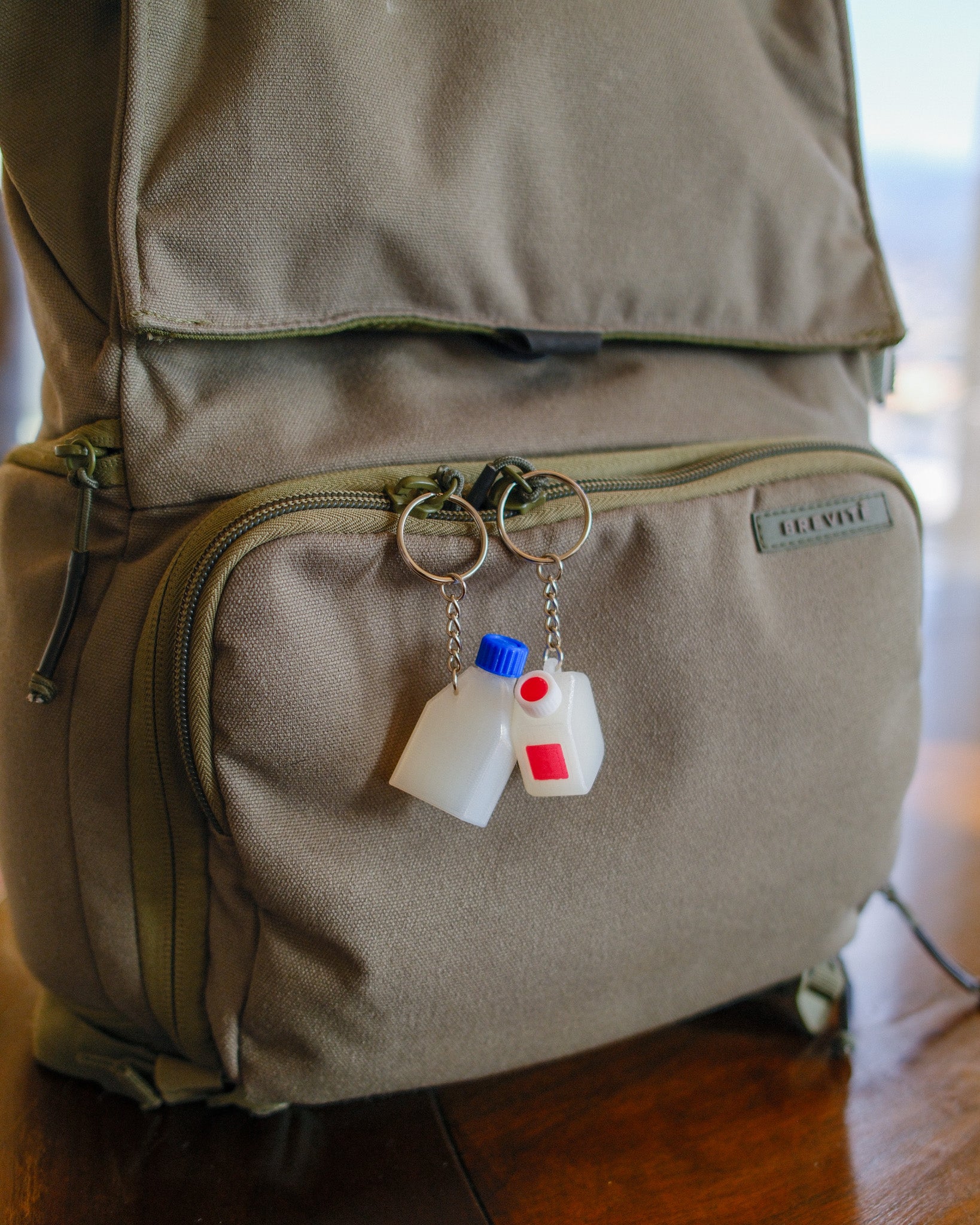 Clear cell culture flask keychain with a blue cap, with a clear media bottle keychain with a white and red cap, hanging off a green backpack
